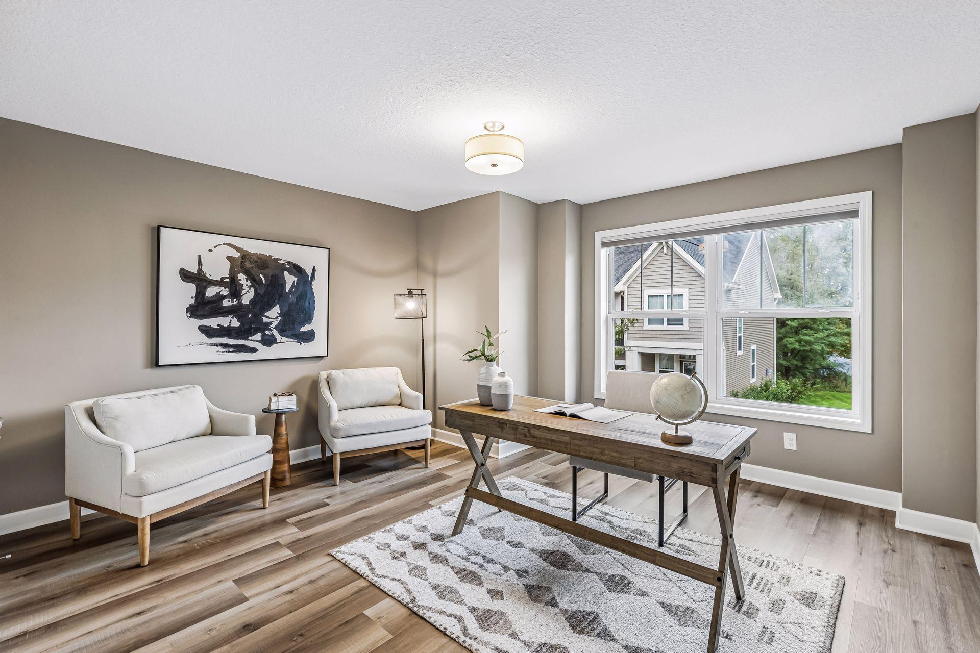 Home office with a wooden desk, white seating, and a large window overlooking greenery.