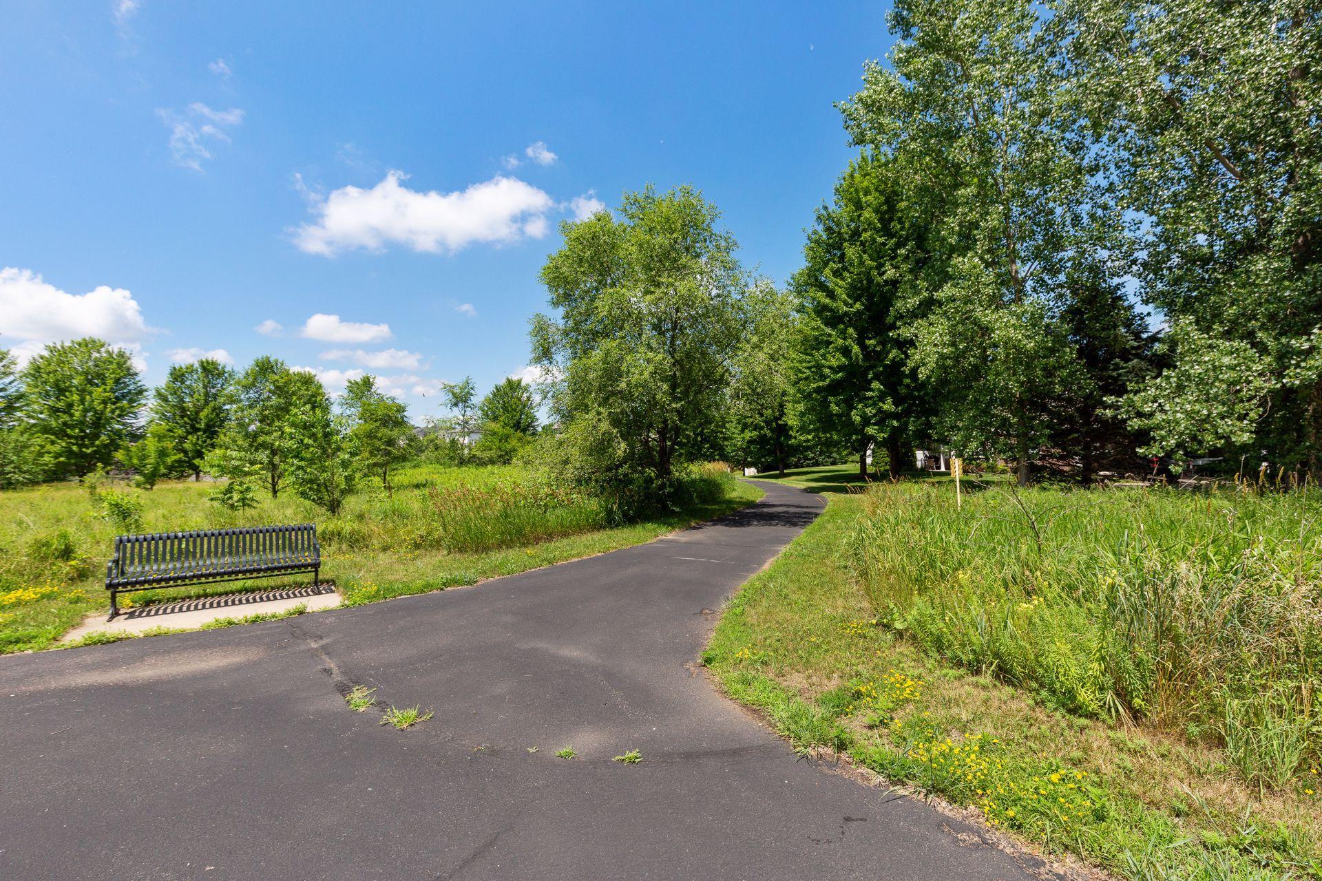 Curving paved park path bordered by trees and grass, with a black bench alongside.