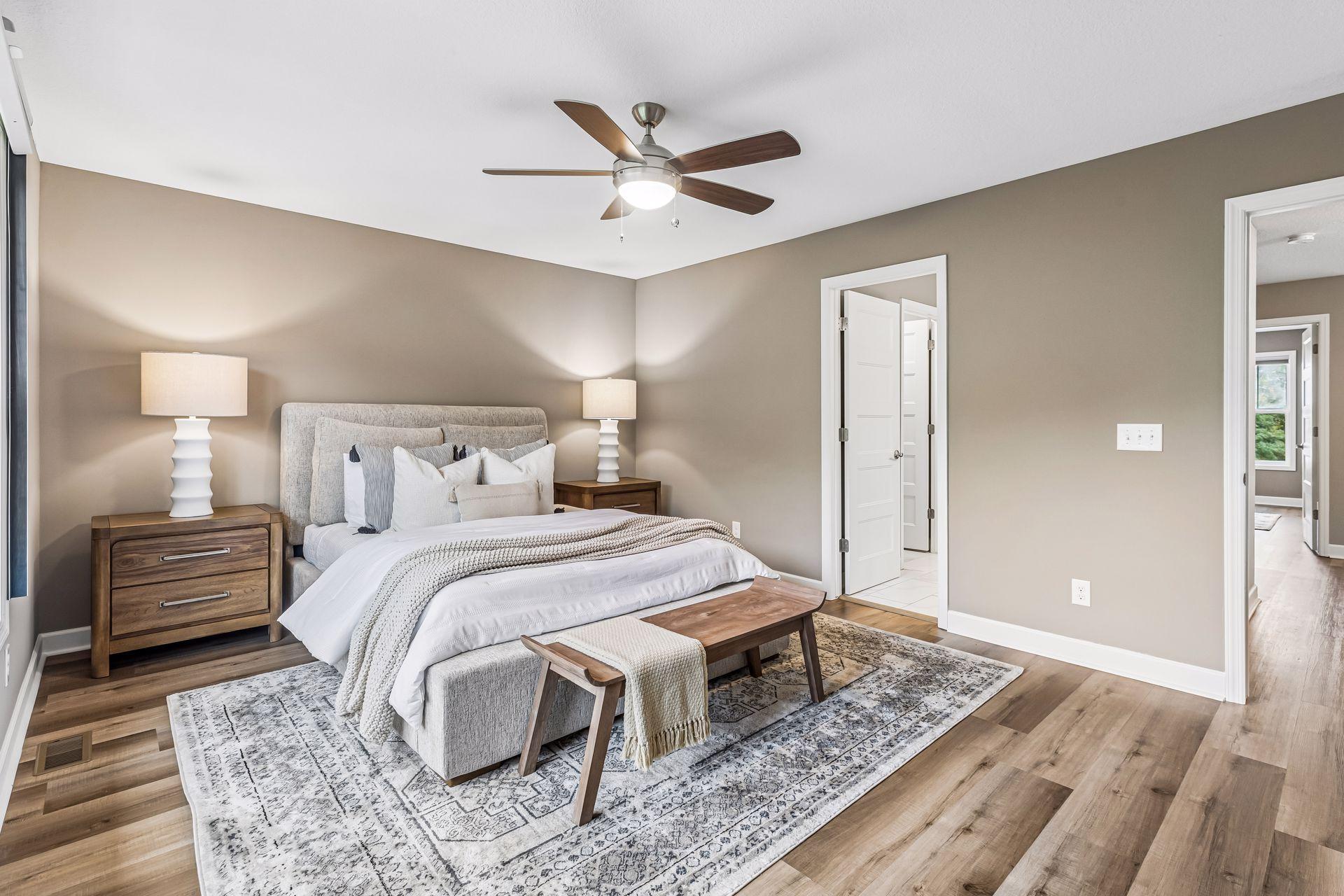 Staged bedroom with white bedding, gray upholstered headboard, wood furniture, and a patterned rug.