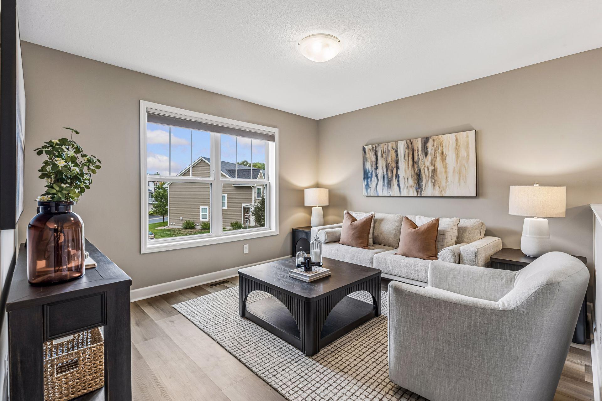 Staged living room with light seating, a dark coffee table, and a large window overlooking nearby homes.