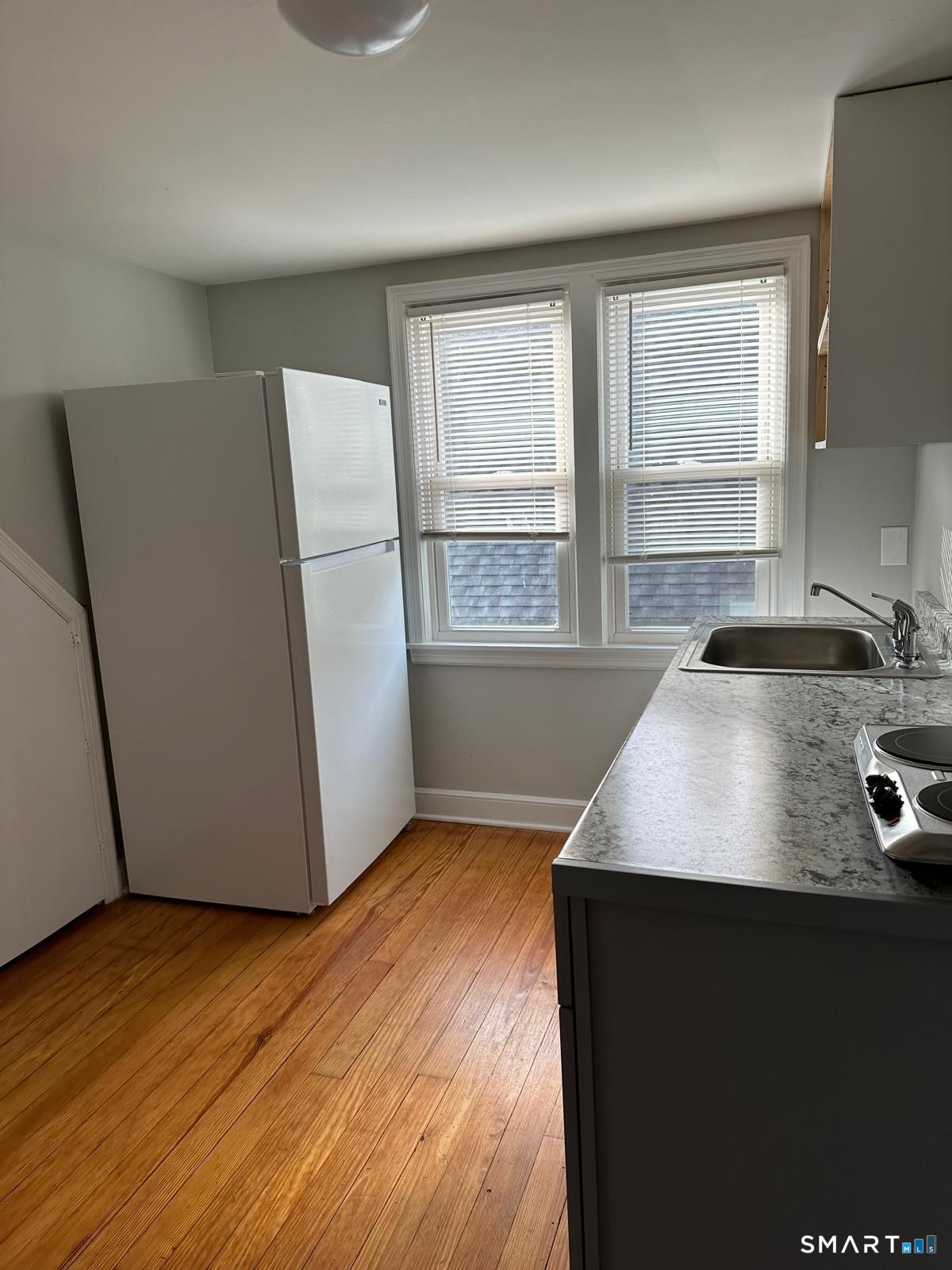 Kitchen with a white refrigerator, gray countertop, two windows, and hardwood floors.