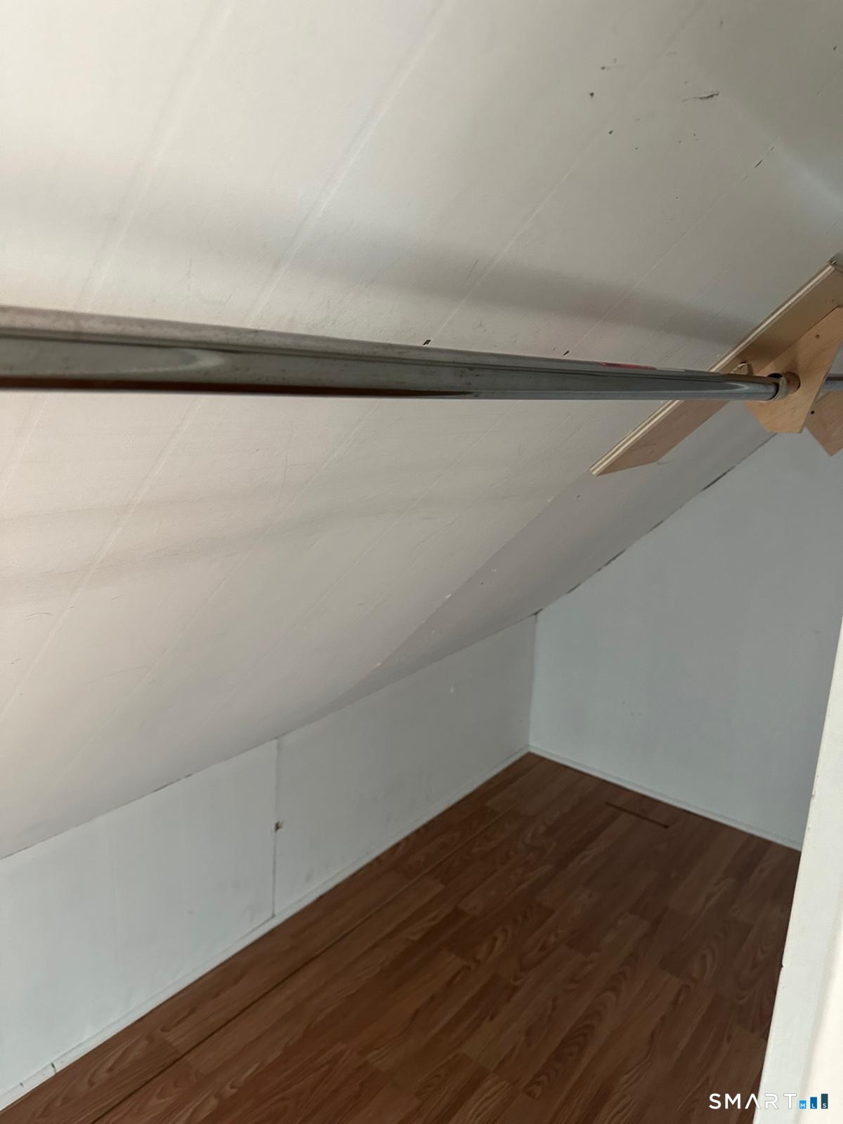 Vacant attic storage nook with a sloped ceiling, laminate floor, and metal hanging rod.