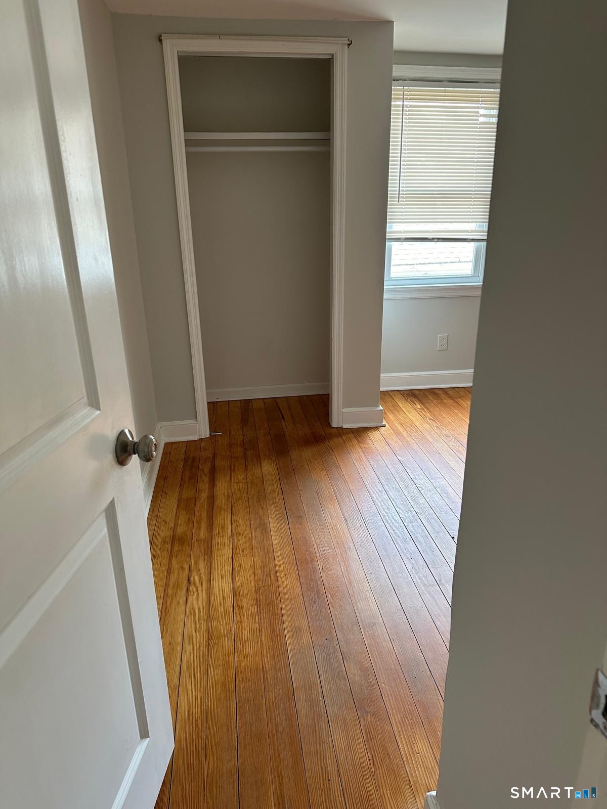 Empty bedroom with a built-in closet, window blinds, and polished hardwood floors.