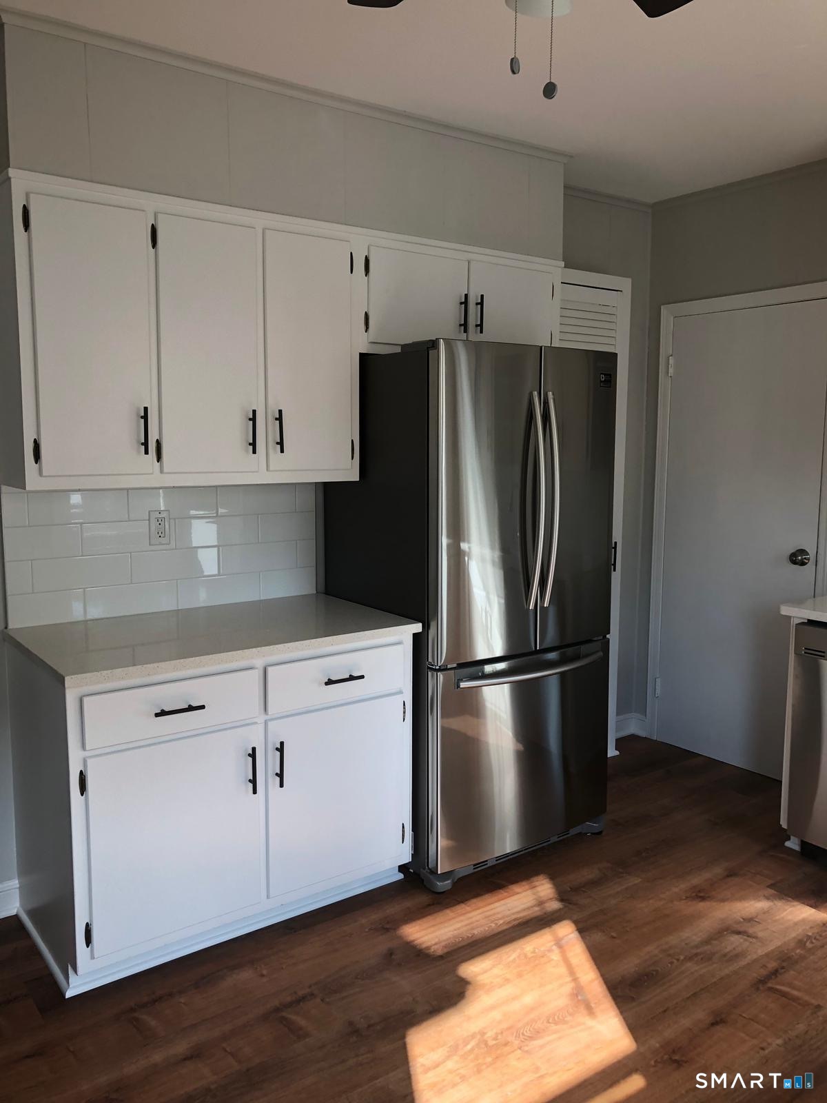 Kitchen with white cabinetry, subway tile backsplash, stainless steel refrigerator, and sunlit wood flooring.