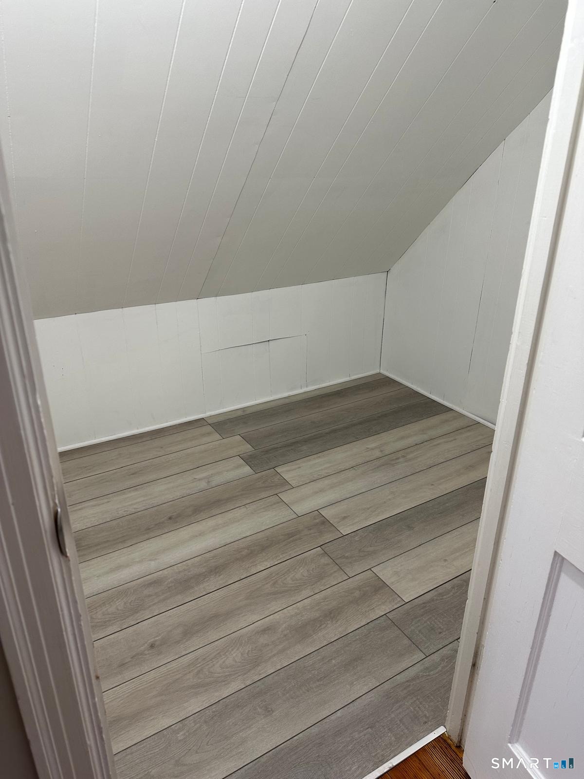 Vacant attic room with a sloped ceiling, white paneling, and gray plank flooring.