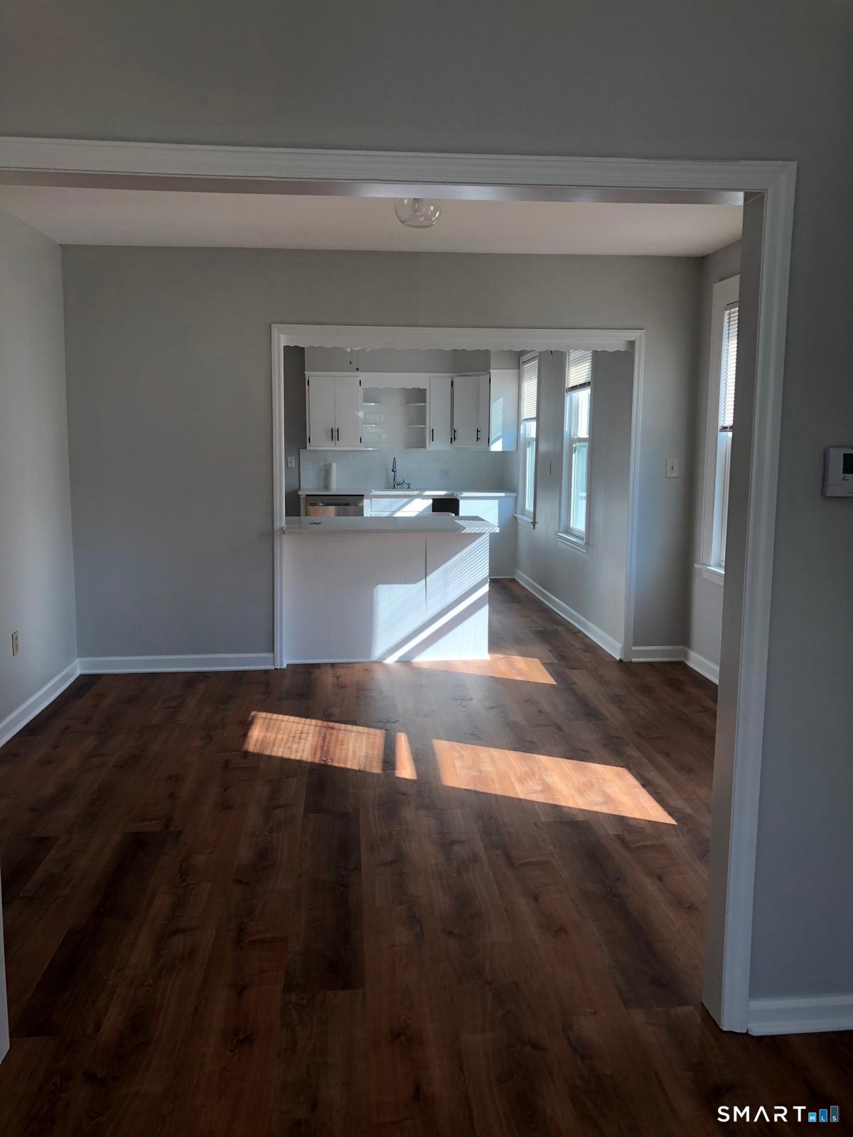 Empty room with dark wood floors opening to a white kitchen with a peninsula and tall windows.