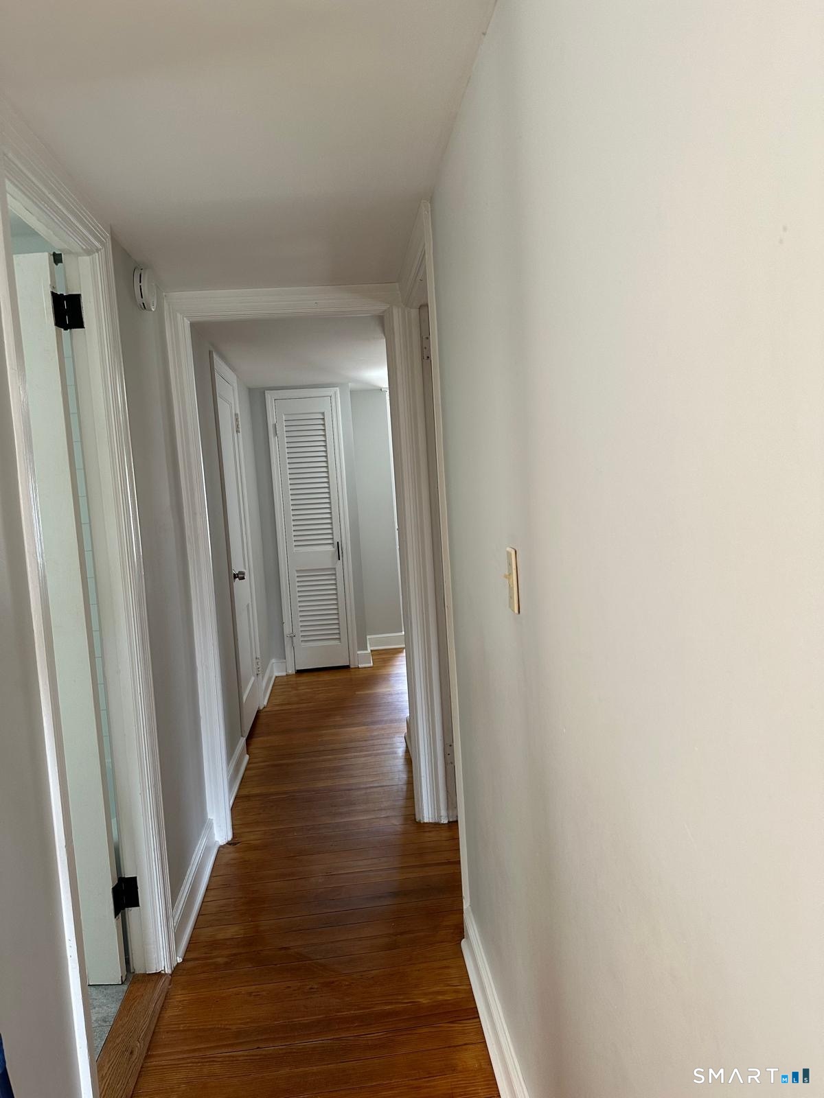 Hallway with hardwood floors, white trim, and a louvered closet door at the far end.