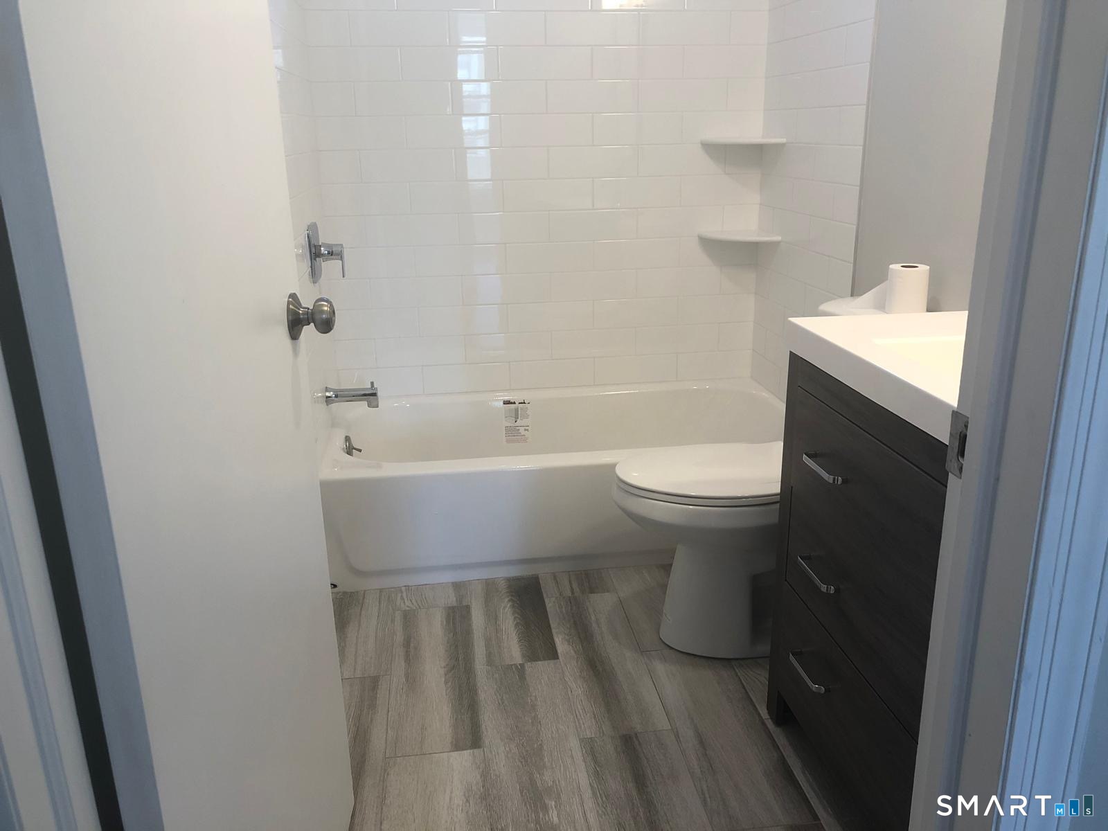 Bathroom with a white subway-tile tub surround, corner shelves, gray tile flooring, and a dark wood vanity.