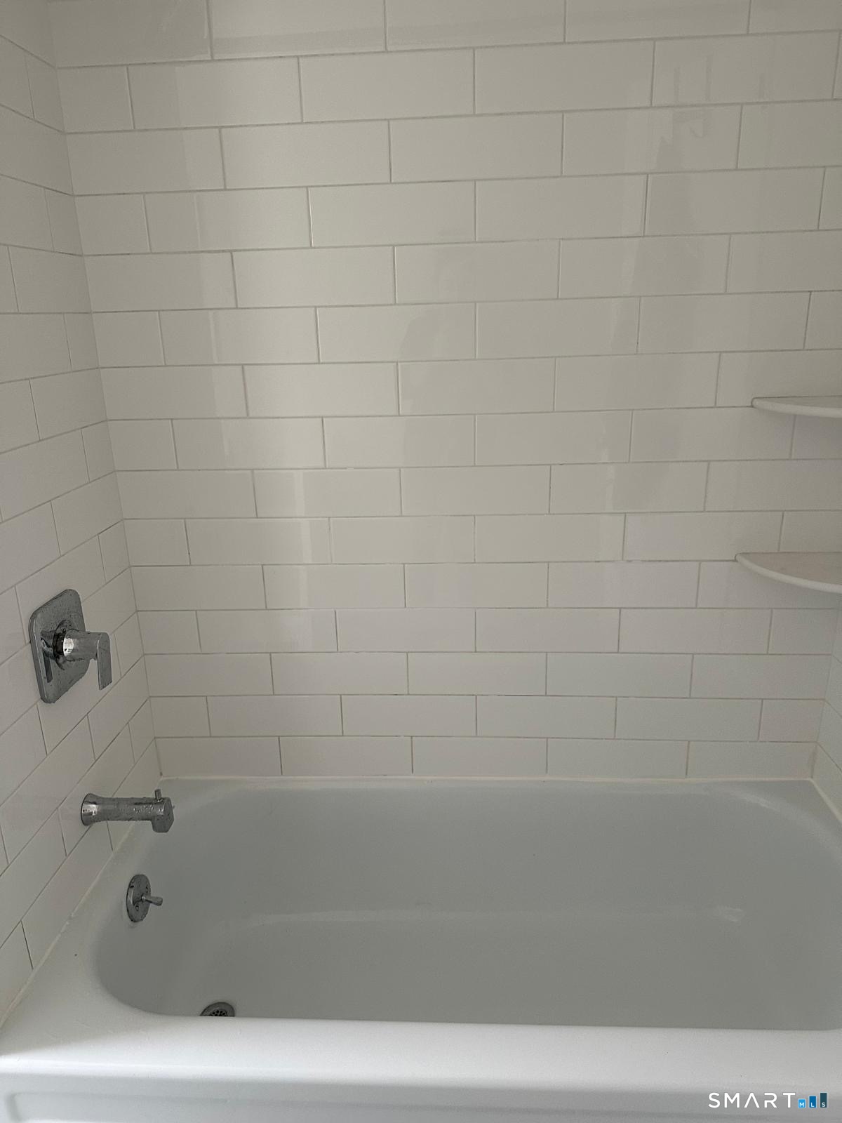 White bathtub with chrome fixtures against white subway tile, with built-in corner shelves.