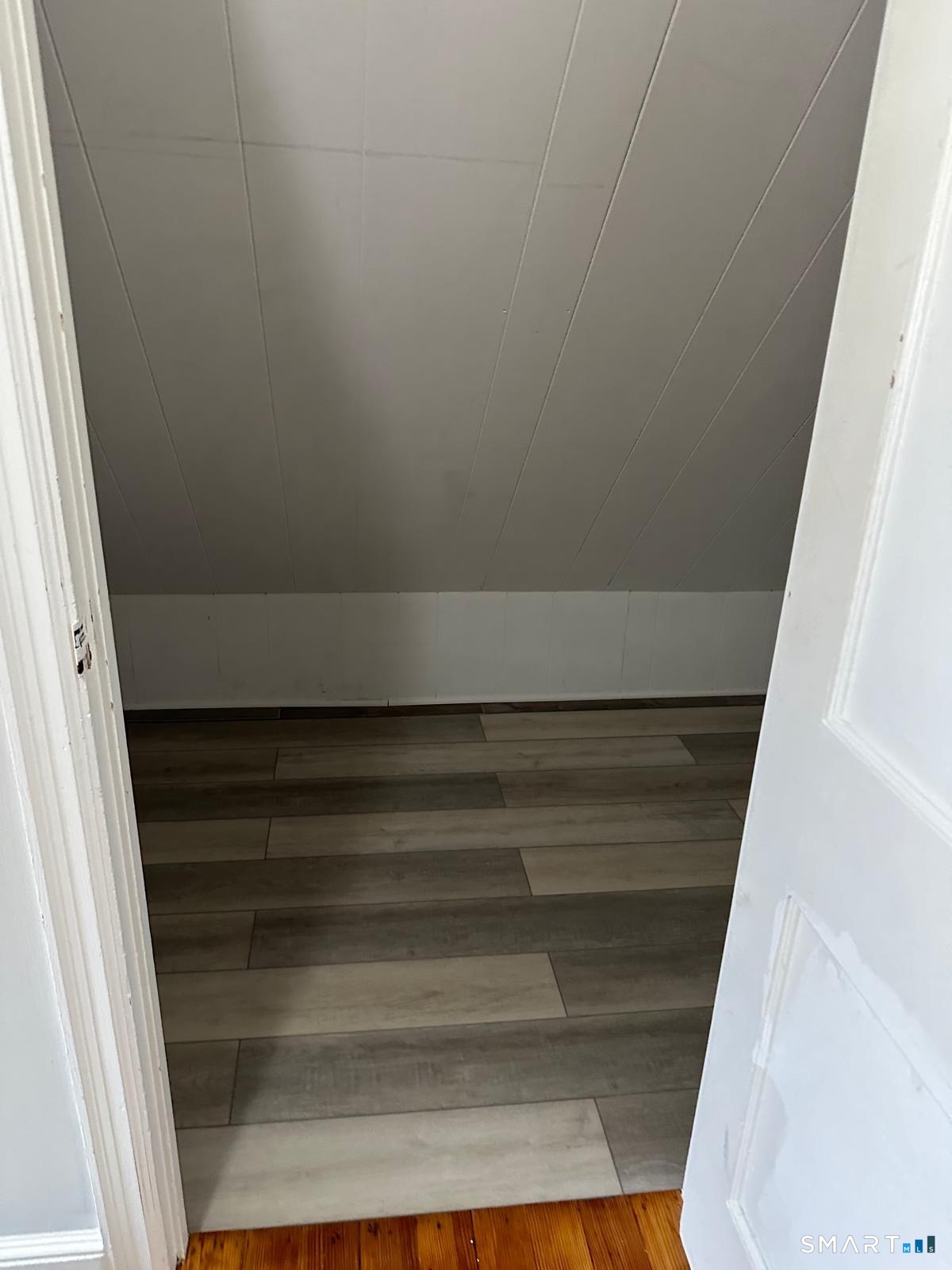 Small vacant room with a sloped ceiling and gray plank flooring, viewed through an open doorway.