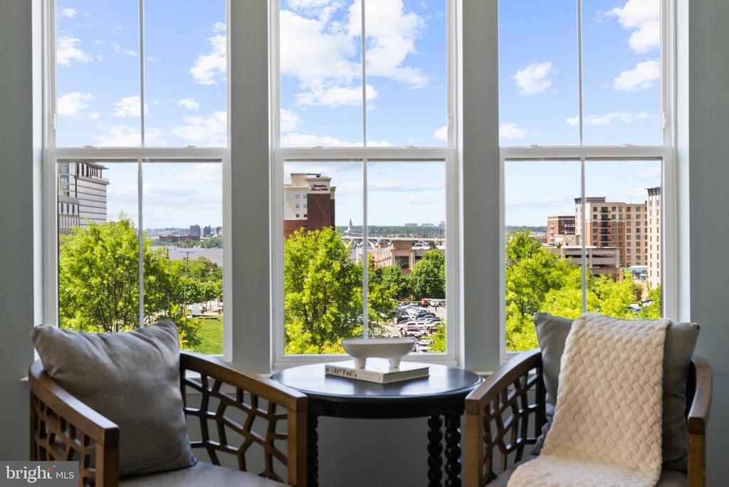 Furnished sitting area with tall windows overlooking trees, buildings, and water.