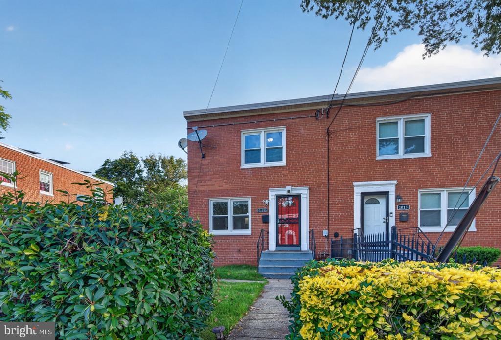 Red-brick rowhouse with a bright red front door, front steps, and leafy shrubs.
