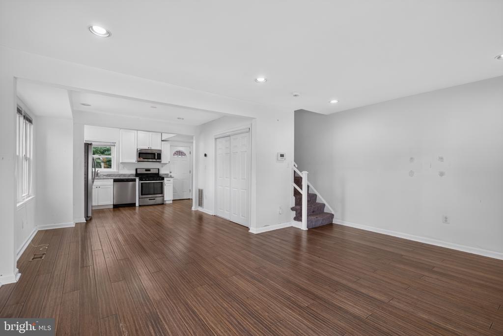 Empty living area with brown plank flooring, open kitchen, and carpeted staircase.