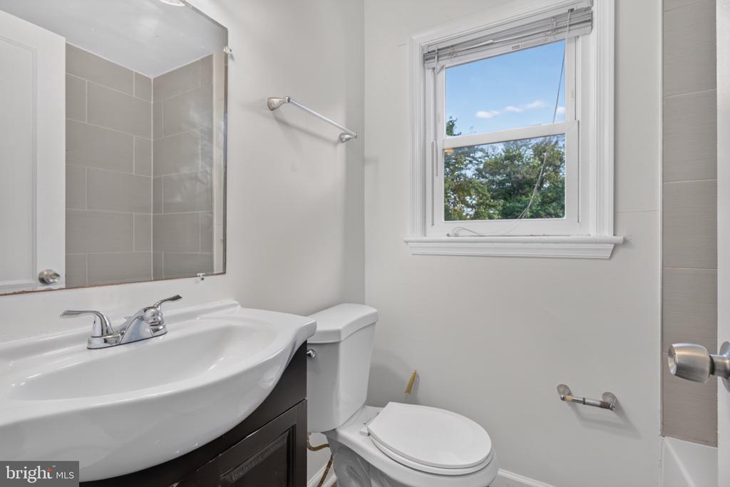 Bathroom with a white sink, toilet, gray-tiled shower, and a window overlooking trees.