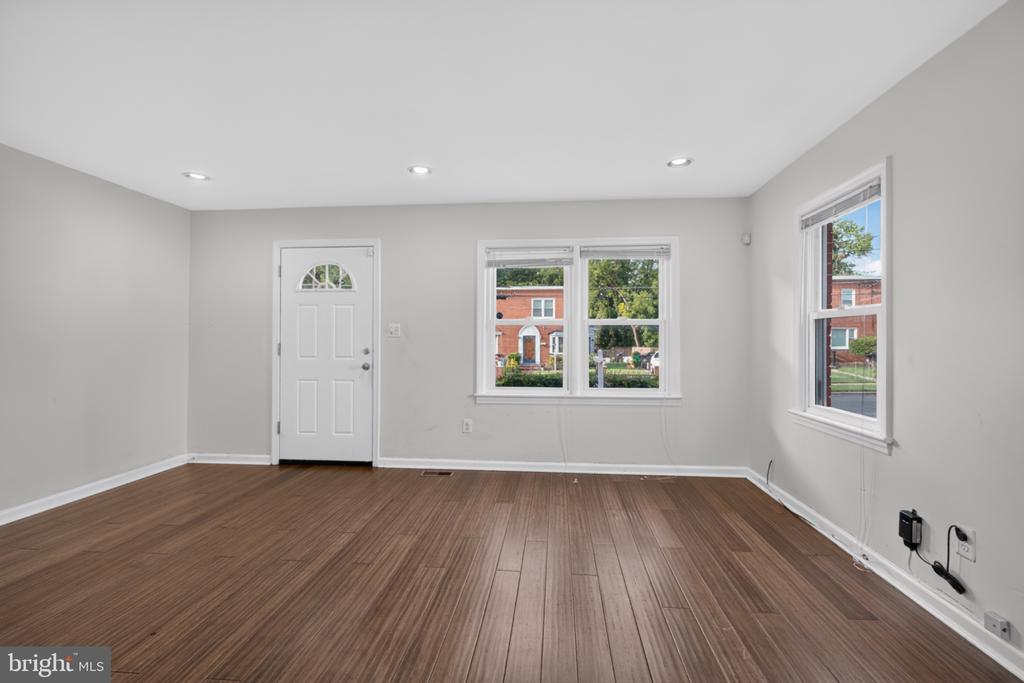 Vacant room with brown plank flooring, recessed lights, a white entry door, and windows overlooking neighboring houses.