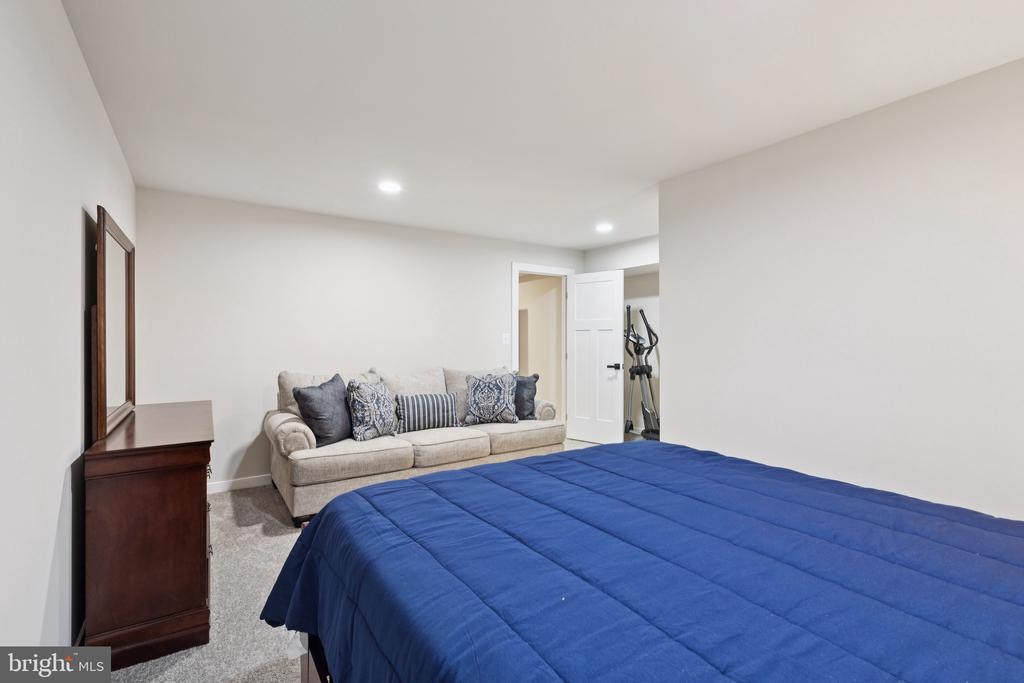 Furnished bedroom with a blue-quilted bed, beige sofa, and exercise machine through the doorway.