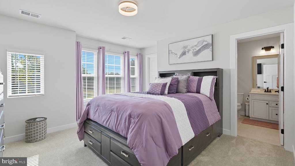 Staged bedroom with purple bedding, a dark wood storage bed, tall windows, and an ensuite bathroom doorway.