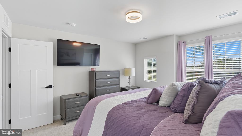 Bedroom with a lavender-and-white bed, gray dressers, wall-mounted TV, and sunlit windows.
