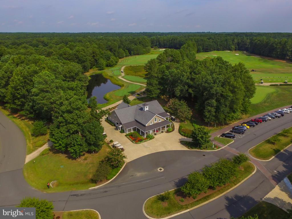 Aerial view of a clubhouse beside golf greens, a pond, and wooded grounds.