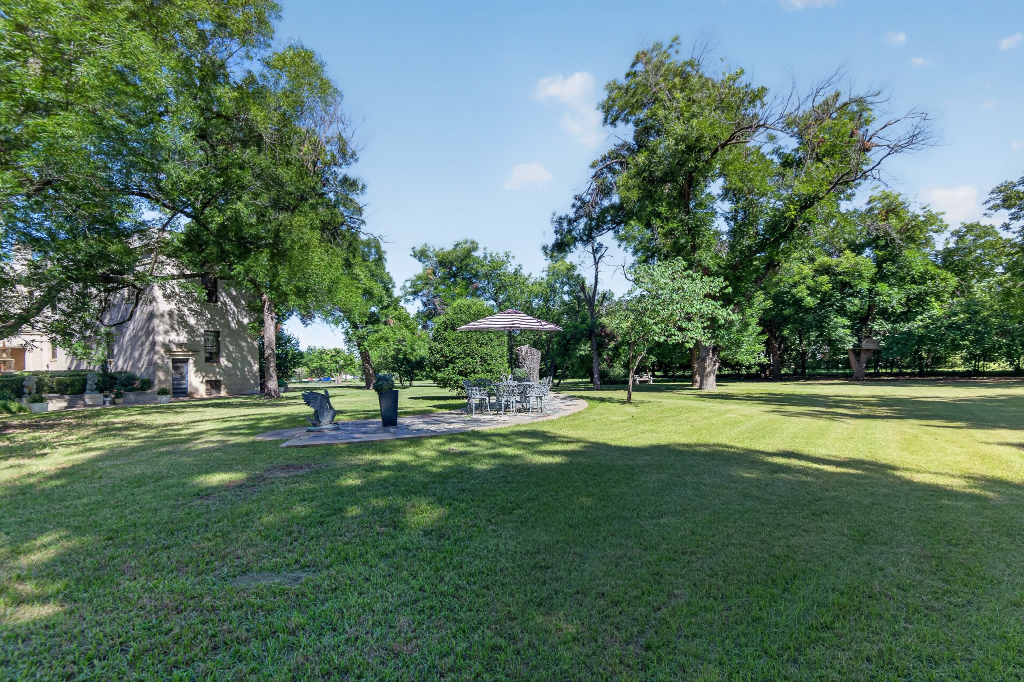 Spacious tree-shaded backyard with a lawn and stone patio dining area beneath an umbrella.