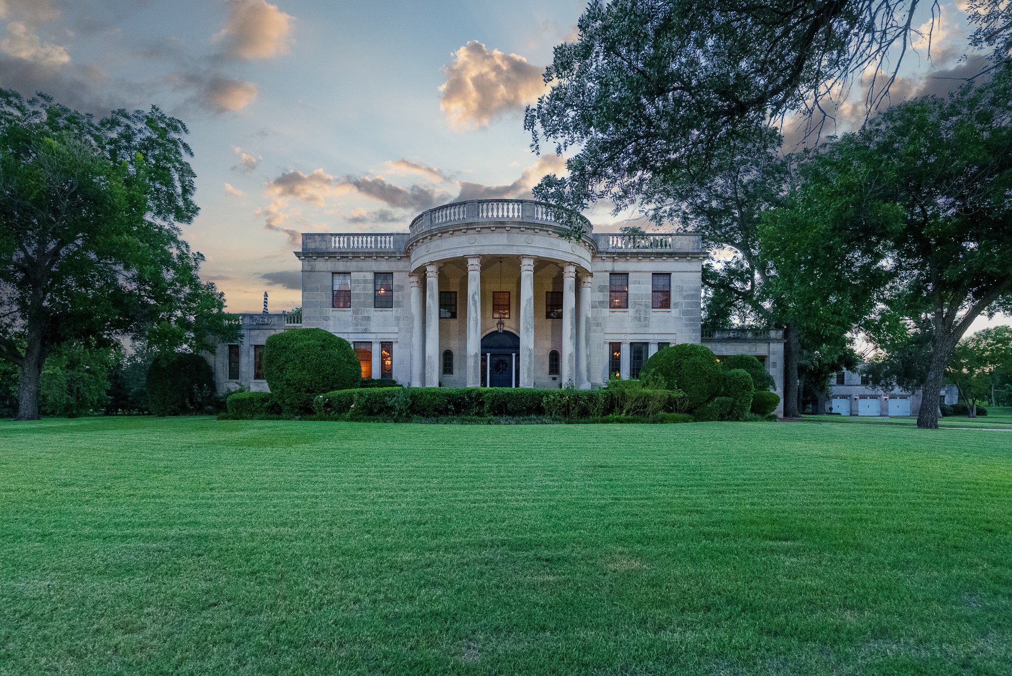 Stone residence with a rounded columned portico and rooftop terrace, framed by trees across a broad lawn.