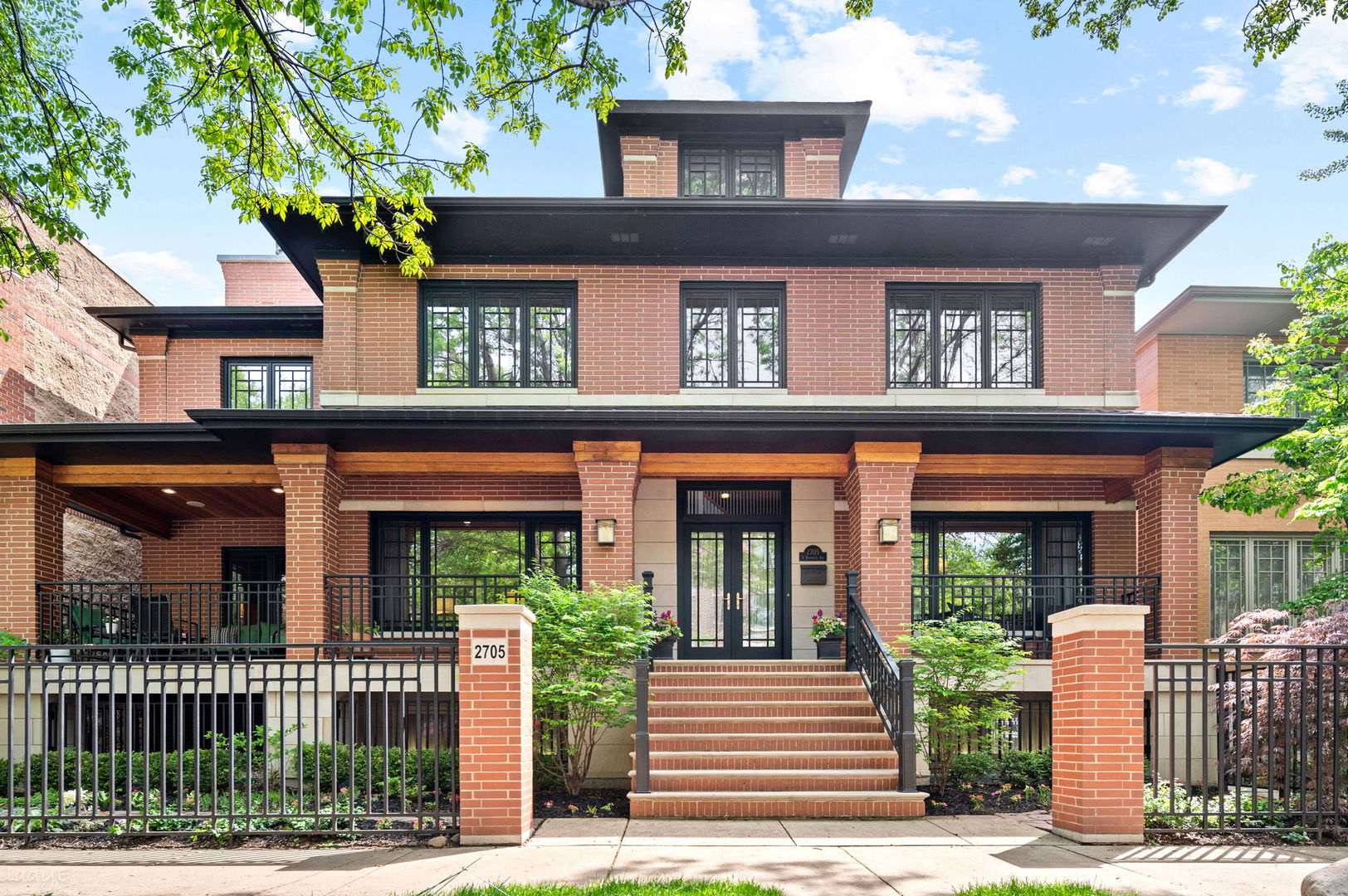 Red-brick home with a broad covered porch, front steps, black railings, and landscaped greenery.