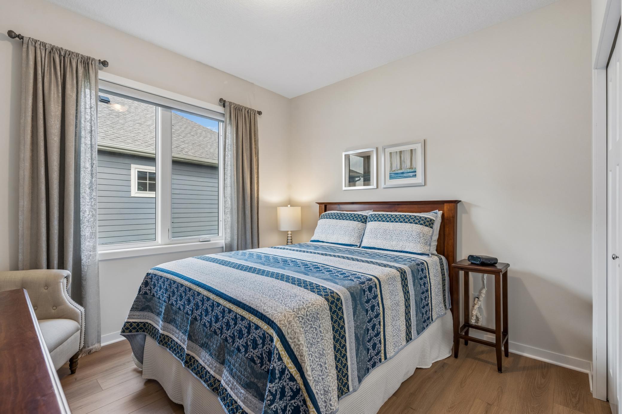 Bedroom with a blue-and-white quilted bed beside a curtained window and upholstered chair.