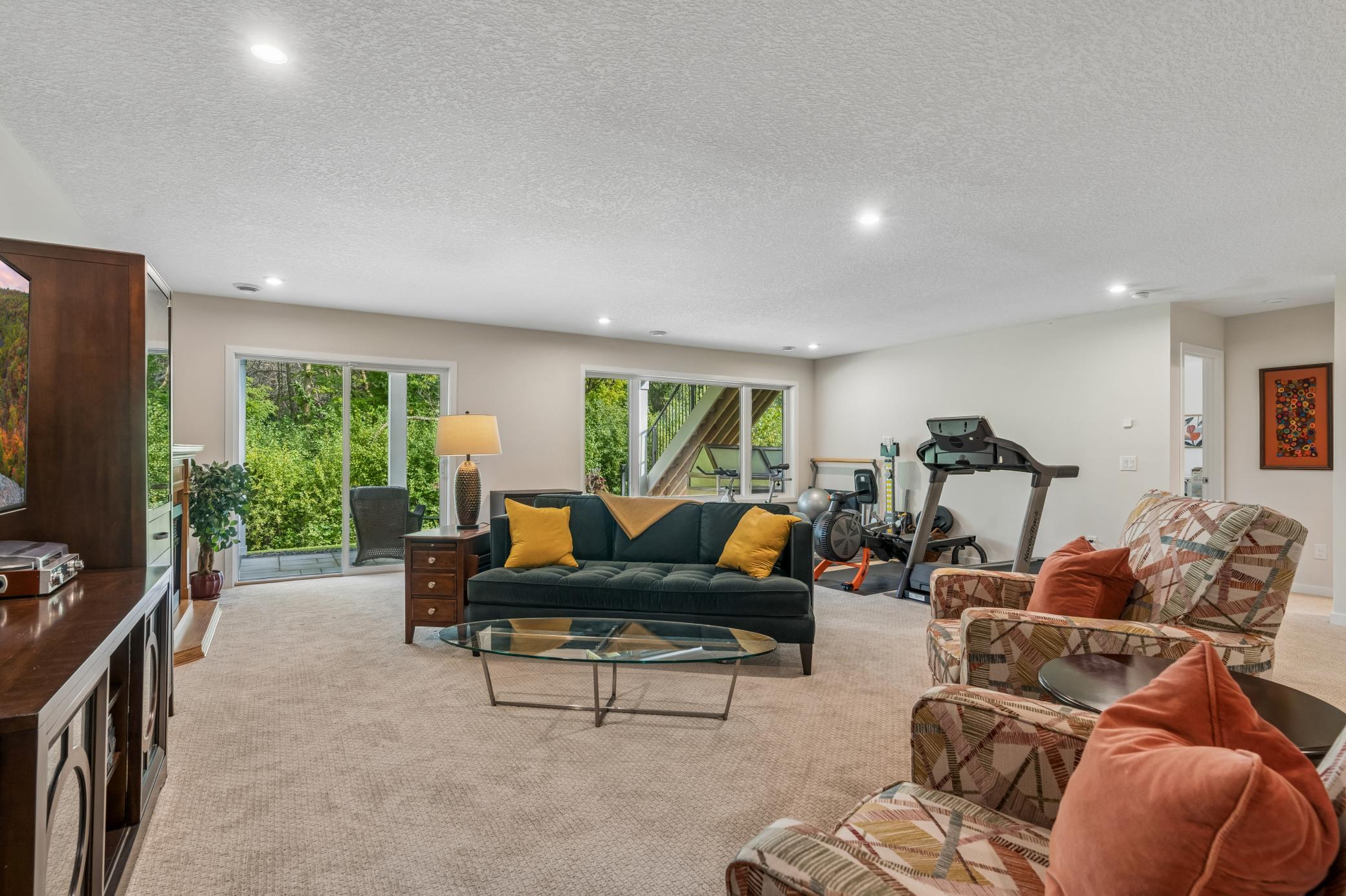 Furnished living room with green sofa, patterned armchairs, large windows, and exercise equipment.