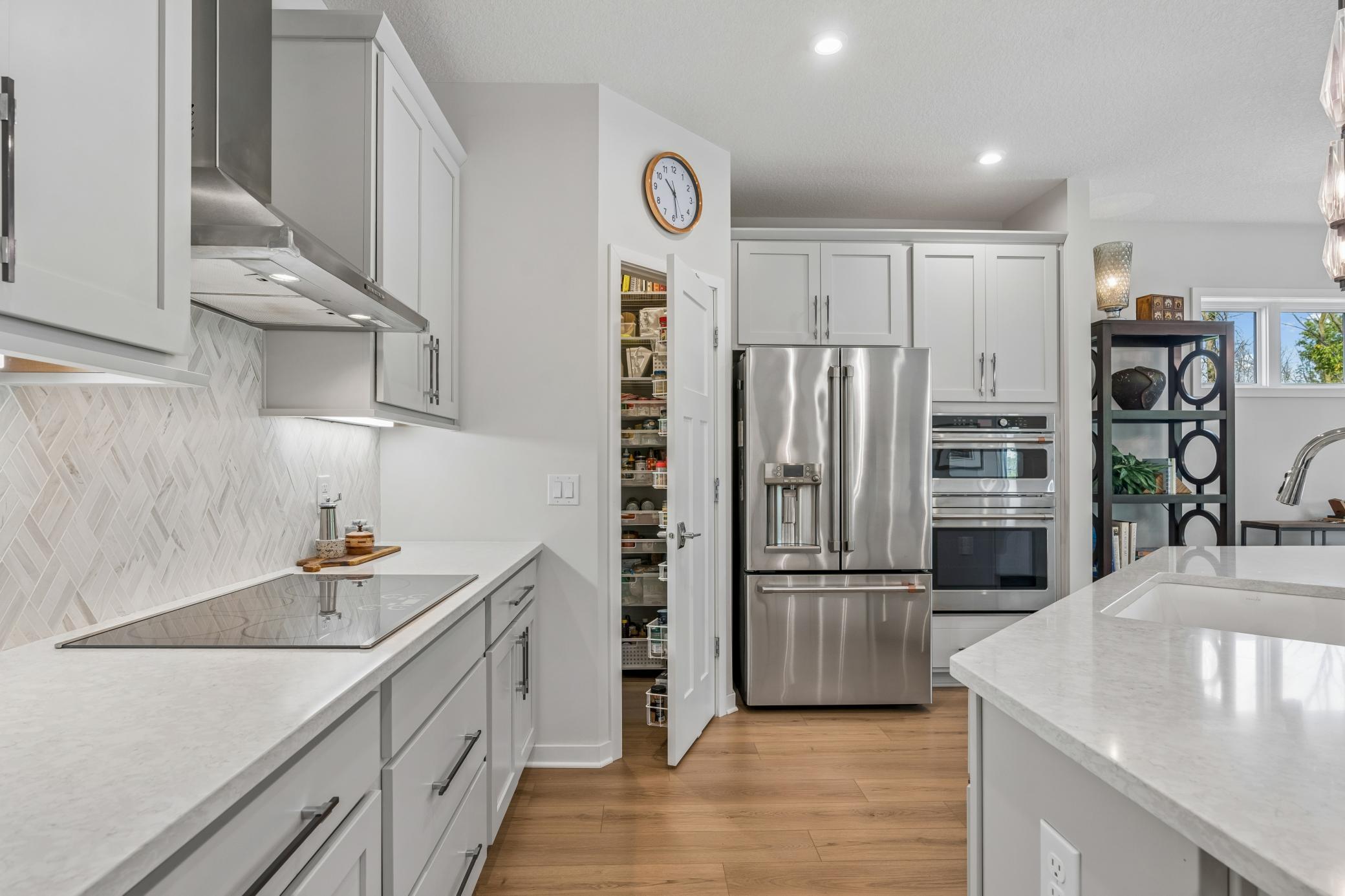 Bright white kitchen with stainless appliances, herringbone backsplash, and an open pantry.