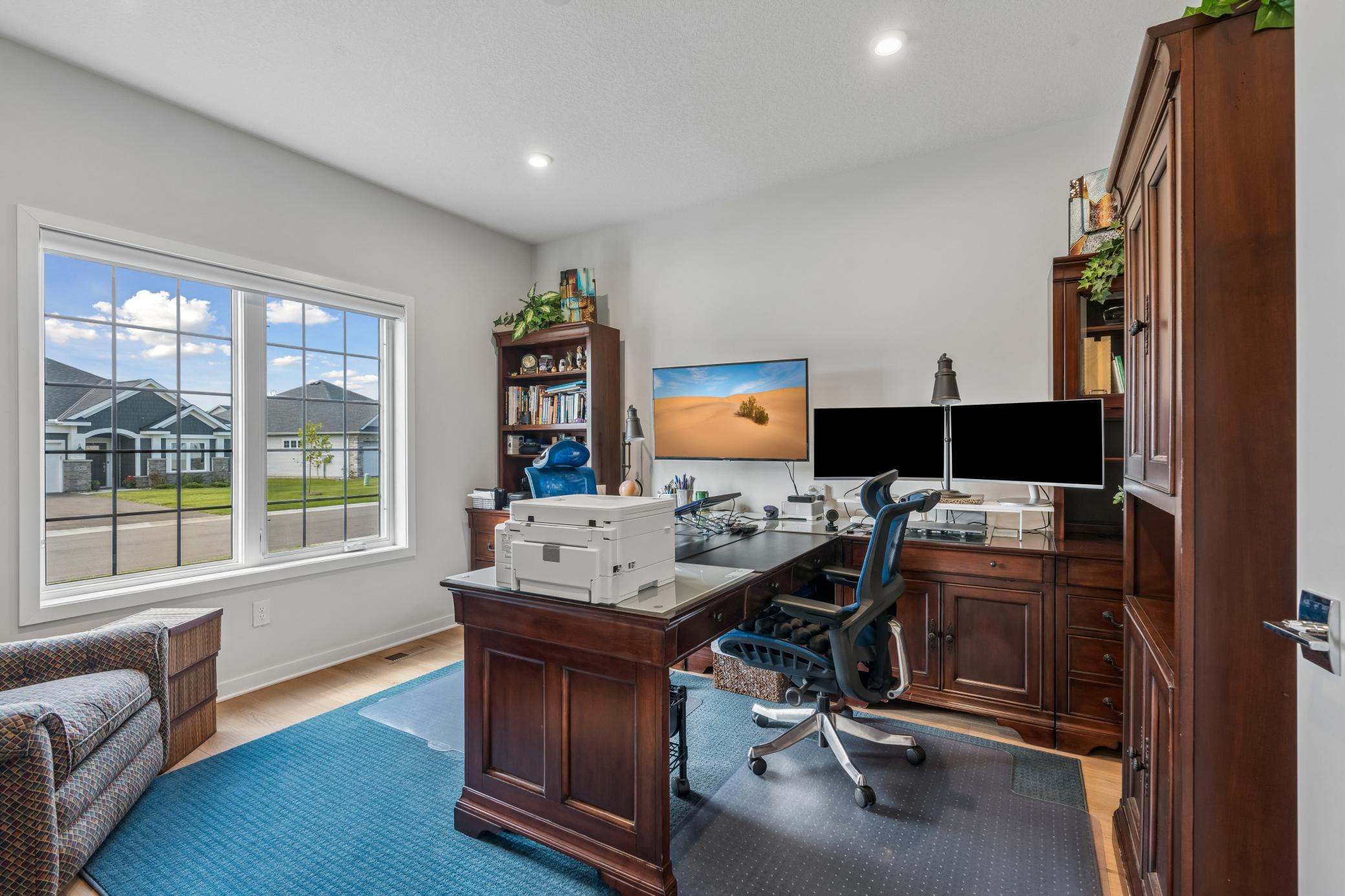 Furnished home office with dark wood cabinetry, dual monitors, and a large window overlooking neighboring homes.