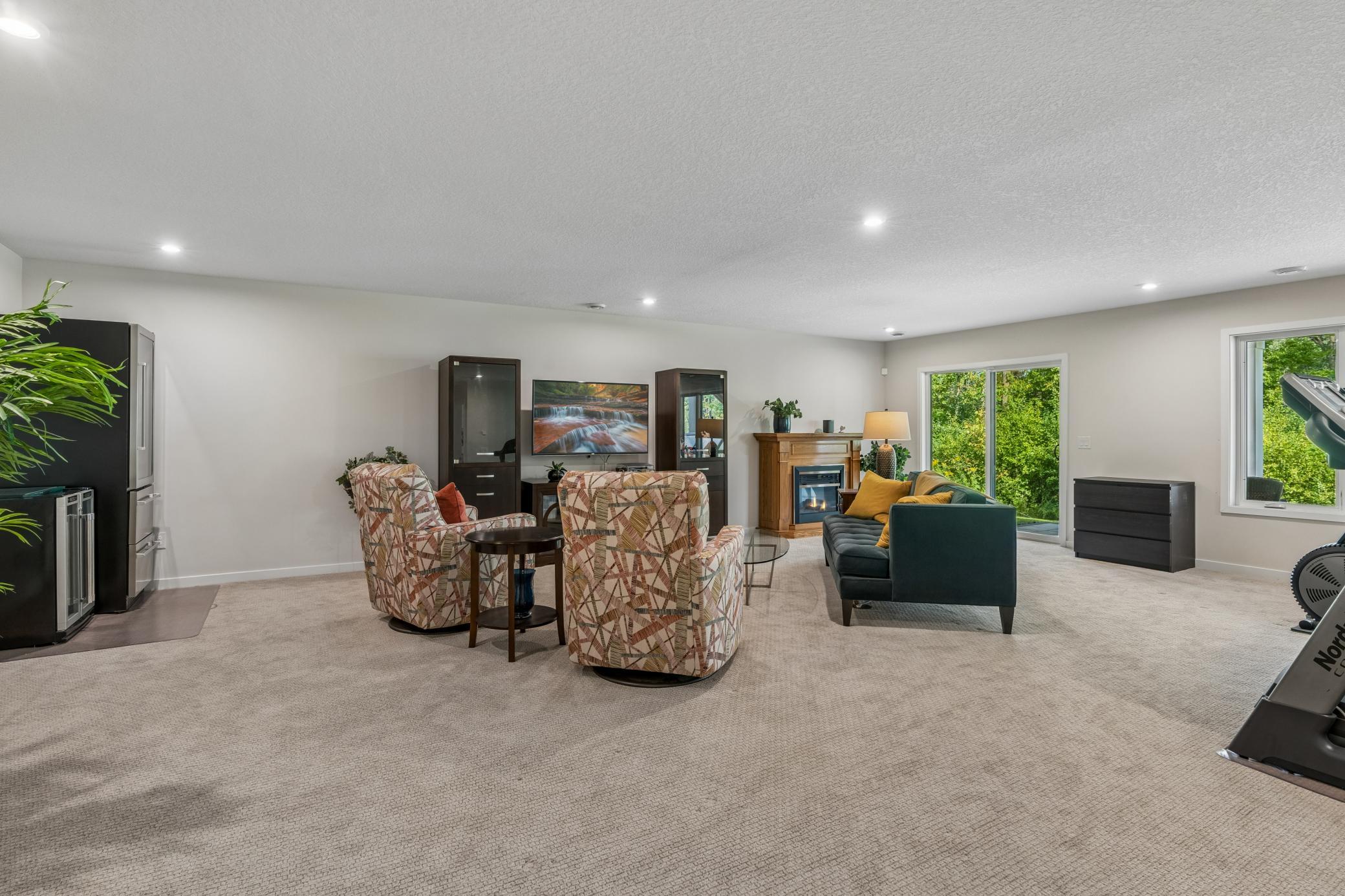 Furnished living room with a fireplace, patterned armchairs, green sofa, and sliding doors overlooking greenery.