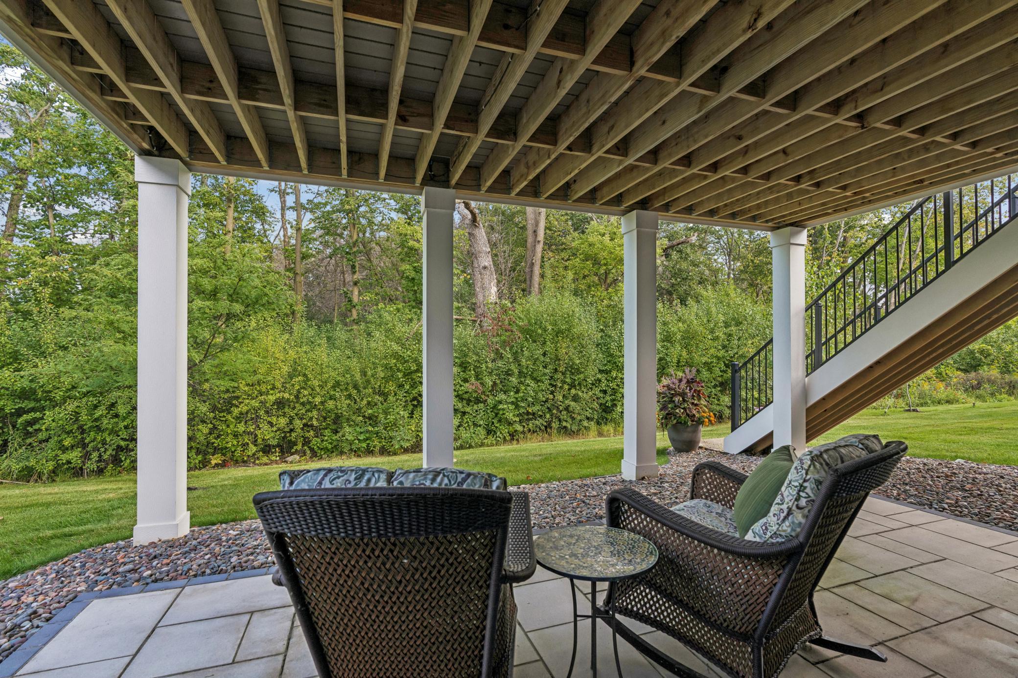 Covered patio with wicker seating beneath a raised deck, overlooking a lawn and trees.