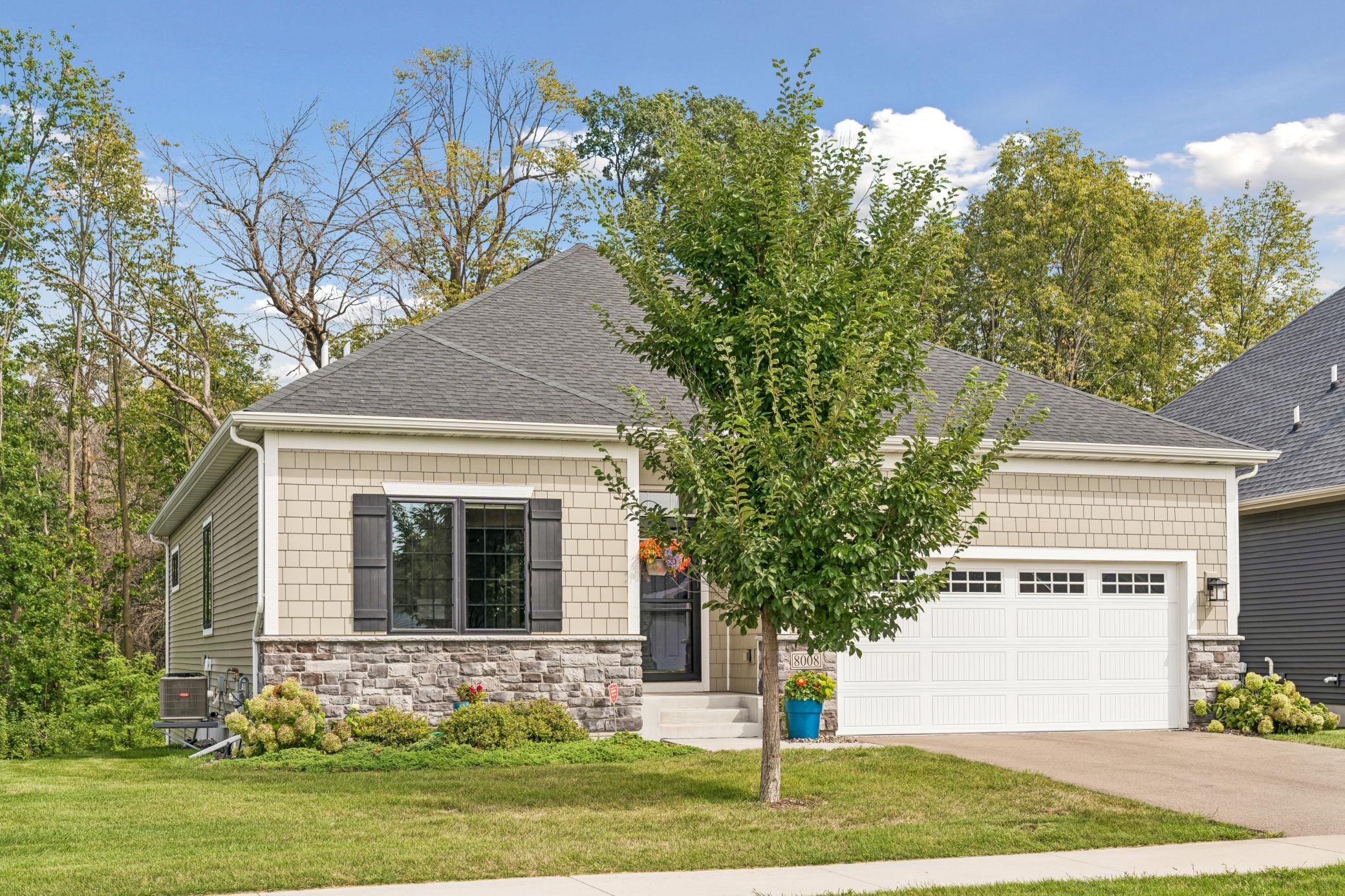 Beige shingle-and-stone house with a white double garage, landscaped lawn, and a tree partly obscuring the entrance.