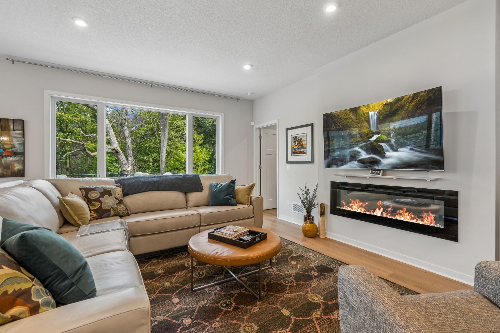 Furnished living room with a sectional sofa, large windows, and a wall-mounted fireplace beneath a television.