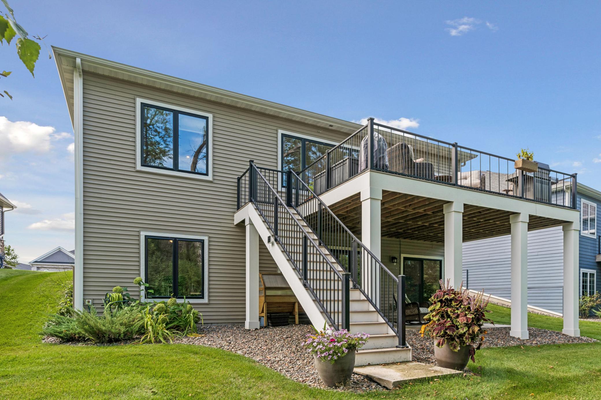 Rear of a tan-sided house with an elevated furnished deck and wide stairs to the landscaped yard.
