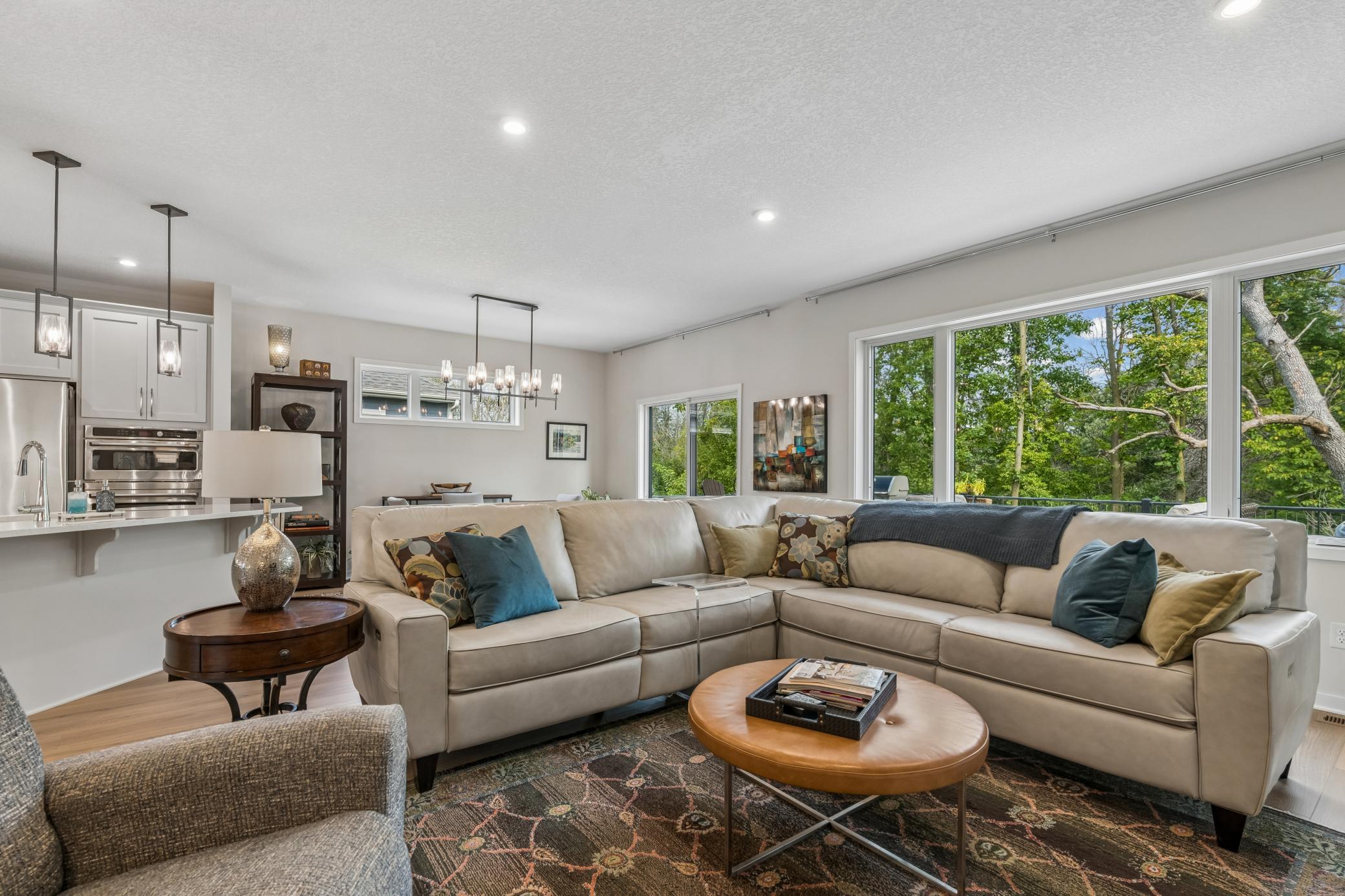 Living room with a beige sectional, open kitchen, and wide windows overlooking leafy trees.