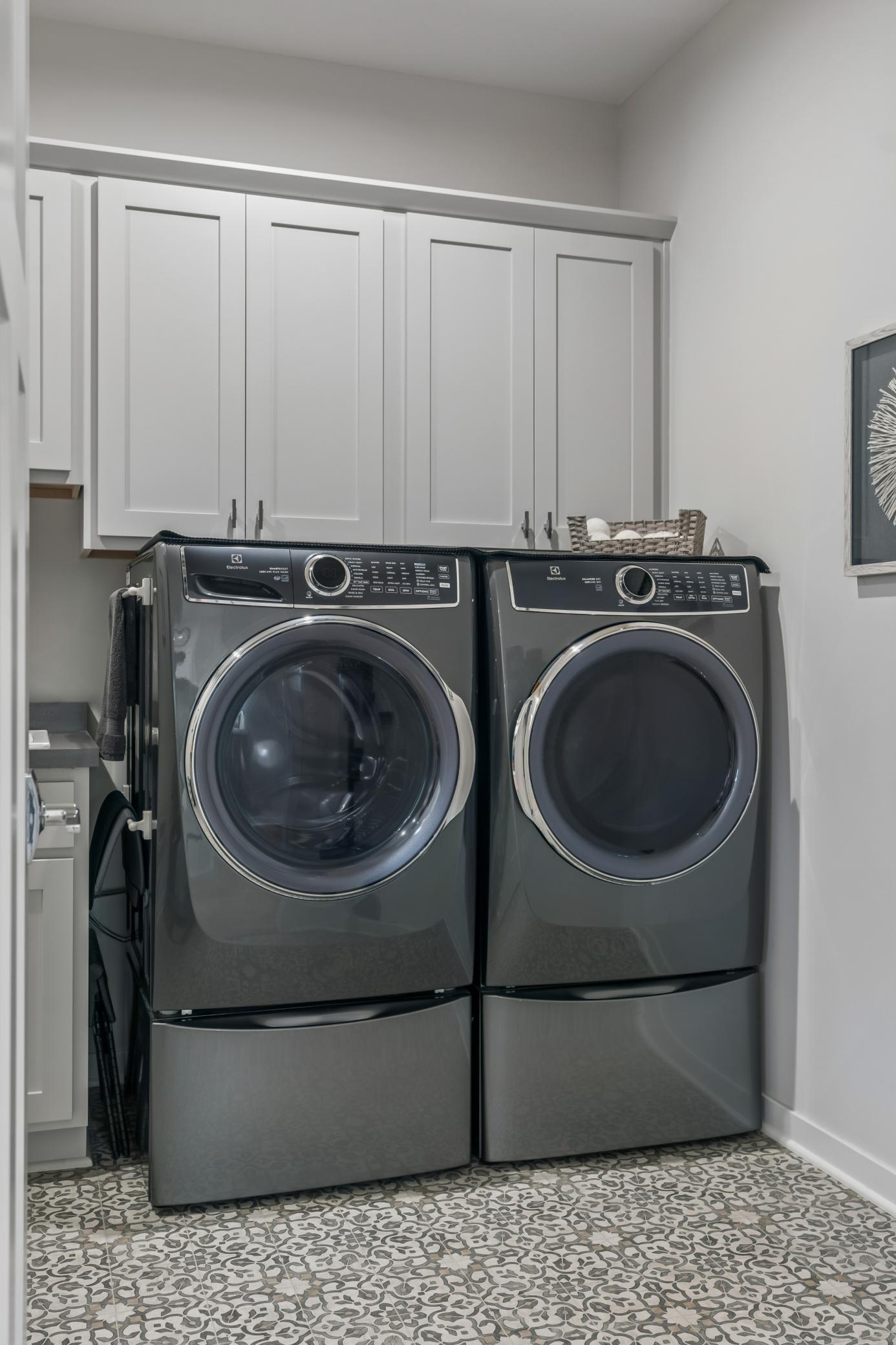 Laundry room with dark gray front-loading washer and dryer, white cabinets, and patterned tile floor.