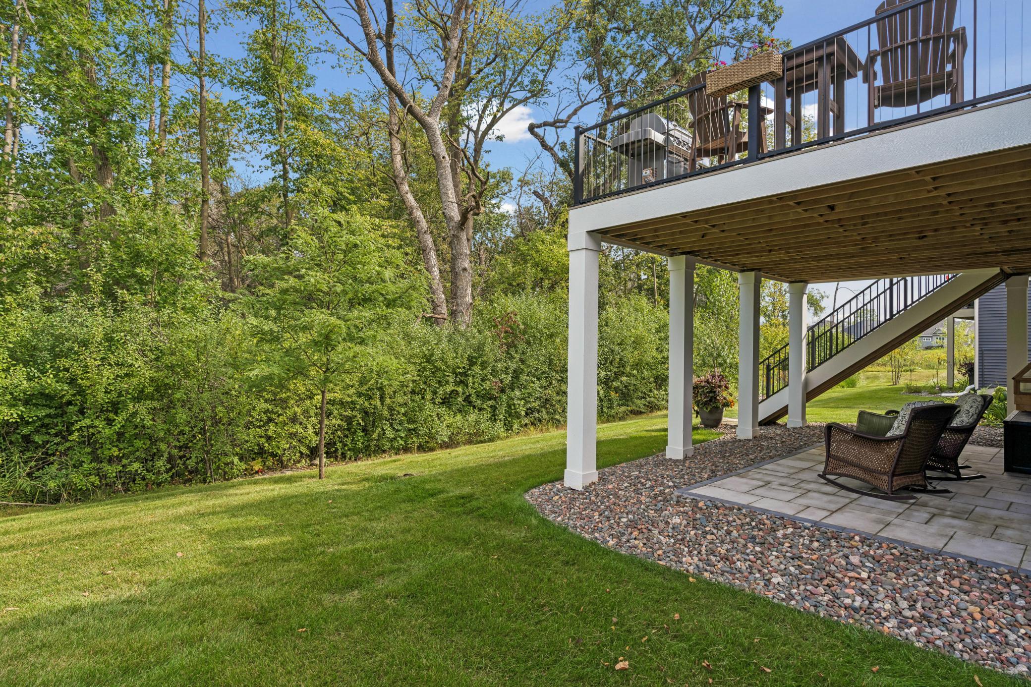 Backyard patio with seating beneath a raised deck, beside a green lawn and mature trees.