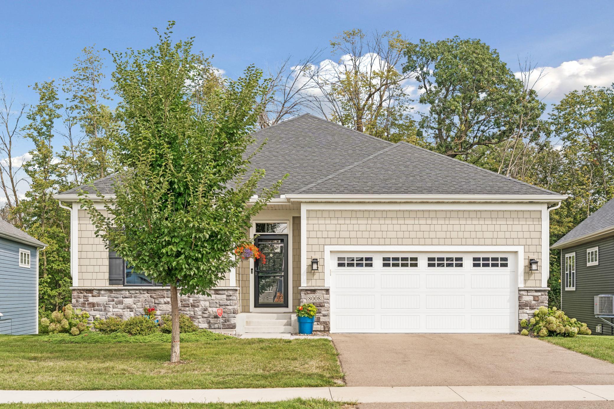 Beige house with stone veneer, a white double garage door, and a leafy tree partly obscuring the front entry.