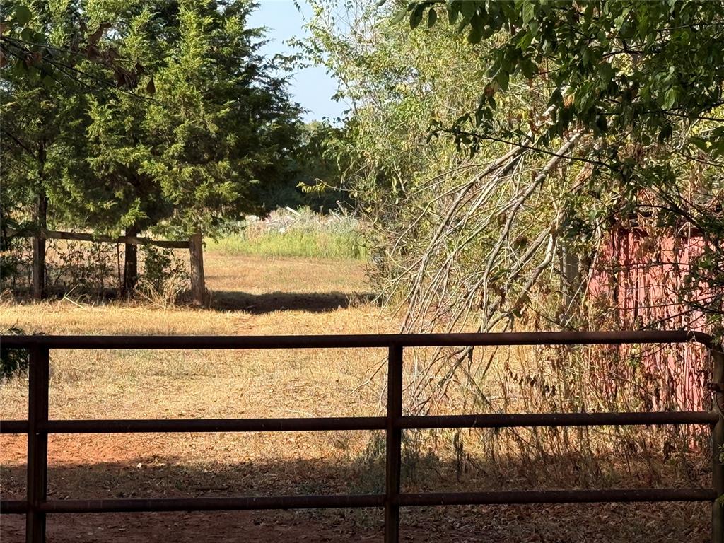 Grassy lot behind a metal gate, shaded by mature trees beside a red outbuilding.