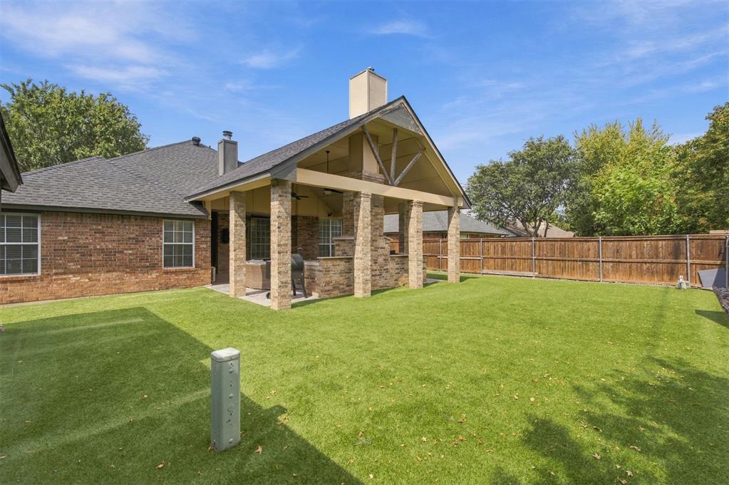 Brick home’s covered backyard patio with stone columns, broad lawn, and wood privacy fence.