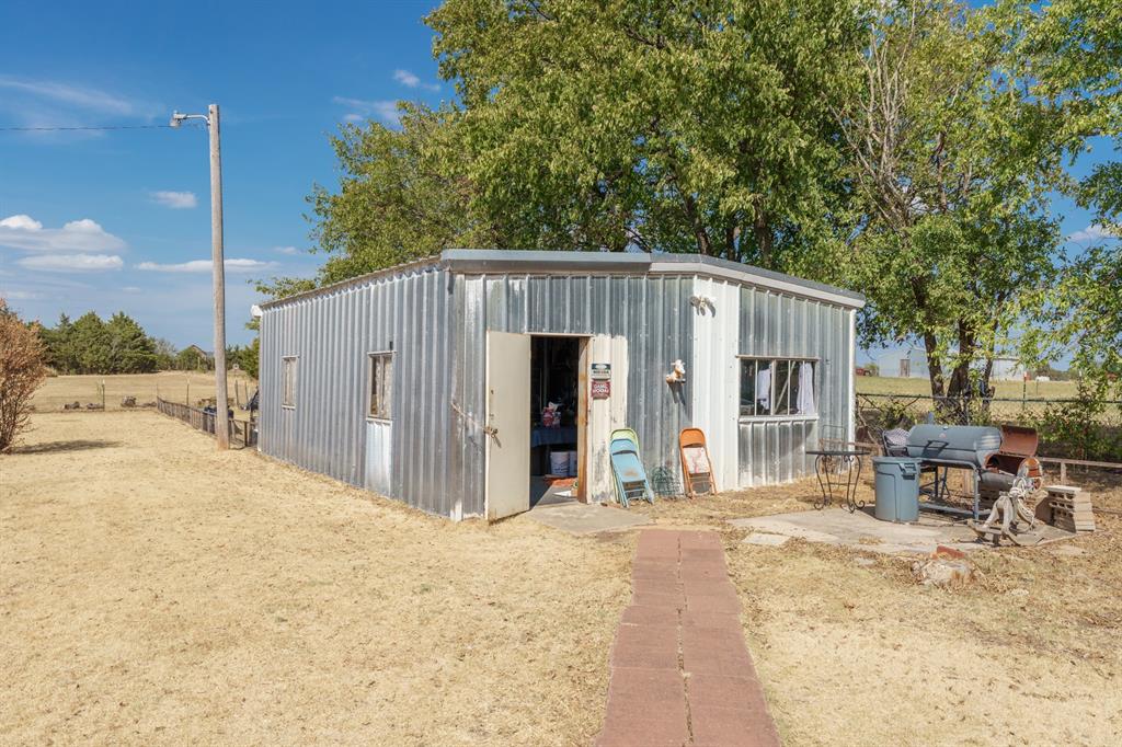 Metal outbuilding with an open door, paved walkway, and outdoor seating beneath a large tree.