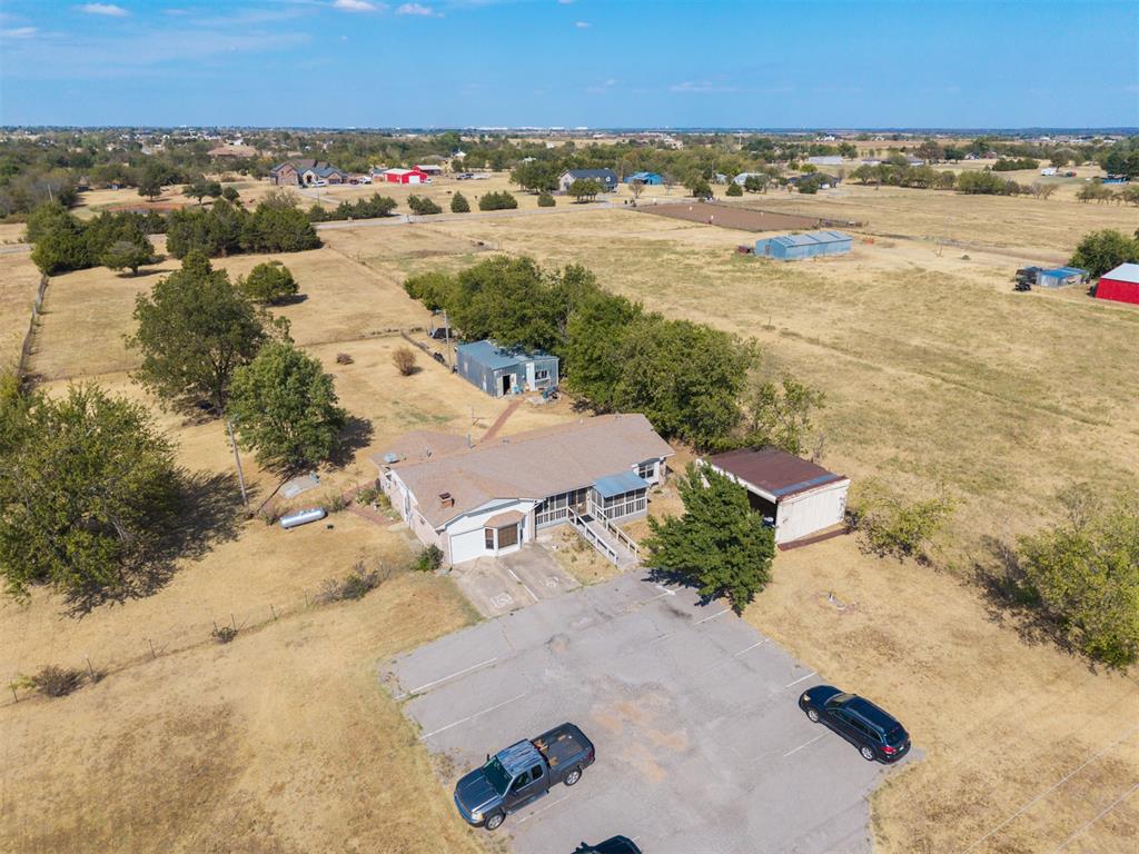 Aerial view of a single-story house with outbuildings, paved parking, and surrounding open fields.