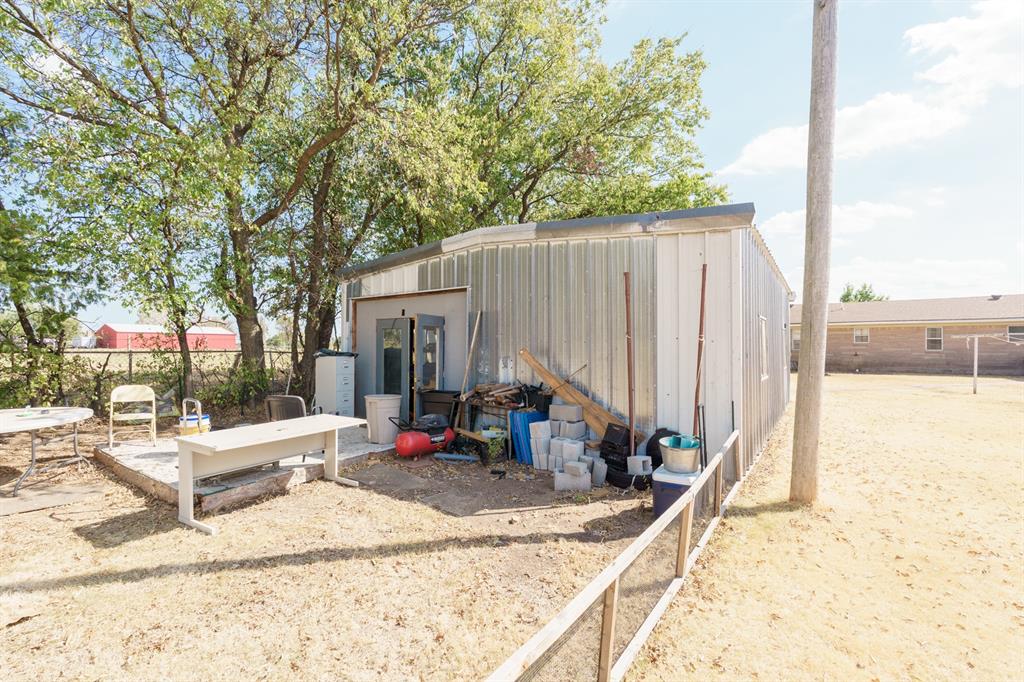 Metal outbuilding with stored equipment beside a dry yard and mature trees.