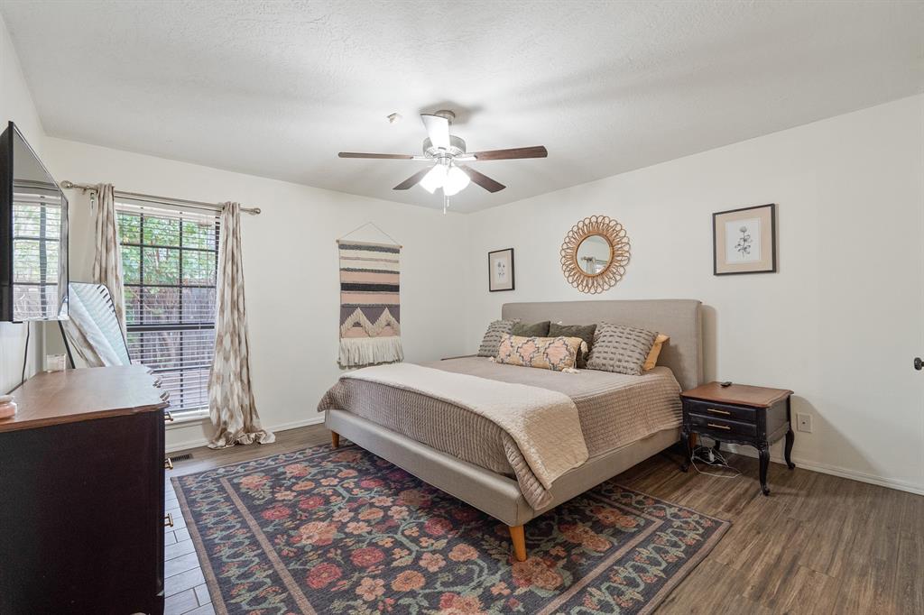 Furnished bedroom with an upholstered bed, colorful patterned rug, and windows with blinds.