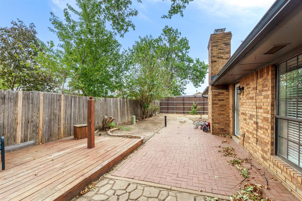 Brick backyard patio beside the home, with a raised wooden deck and mature trees.
