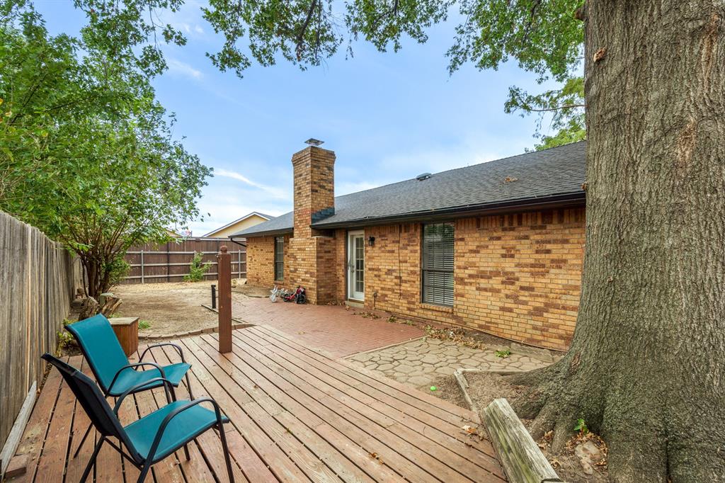 Brick house backyard with a wooden deck, patio chairs, and a large mature tree.