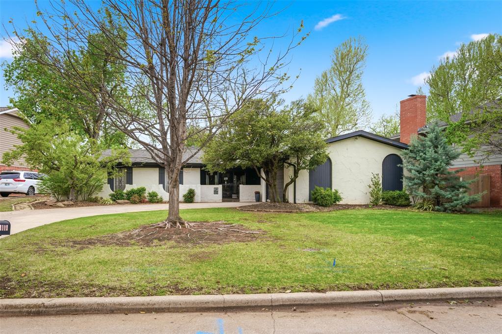 Single-story stucco home with arched windows and a brick chimney behind a lawn and large tree.