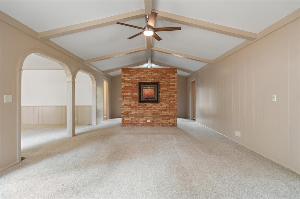 Empty living room with vaulted beamed ceiling, ceiling fan, and central brick wall.