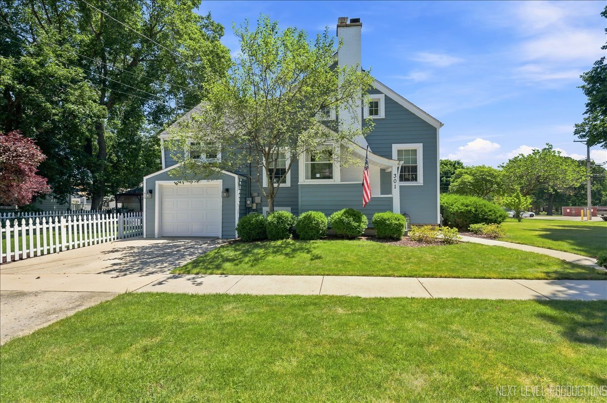 Blue-gray two-story house with an attached garage, white picket fence, and American flag.